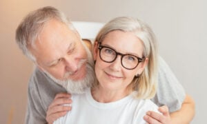 A woman in her late 50s/early 60s smiles, as her husband is behind her about to give her a hug. They're wondering about GICs for his retirement savigs