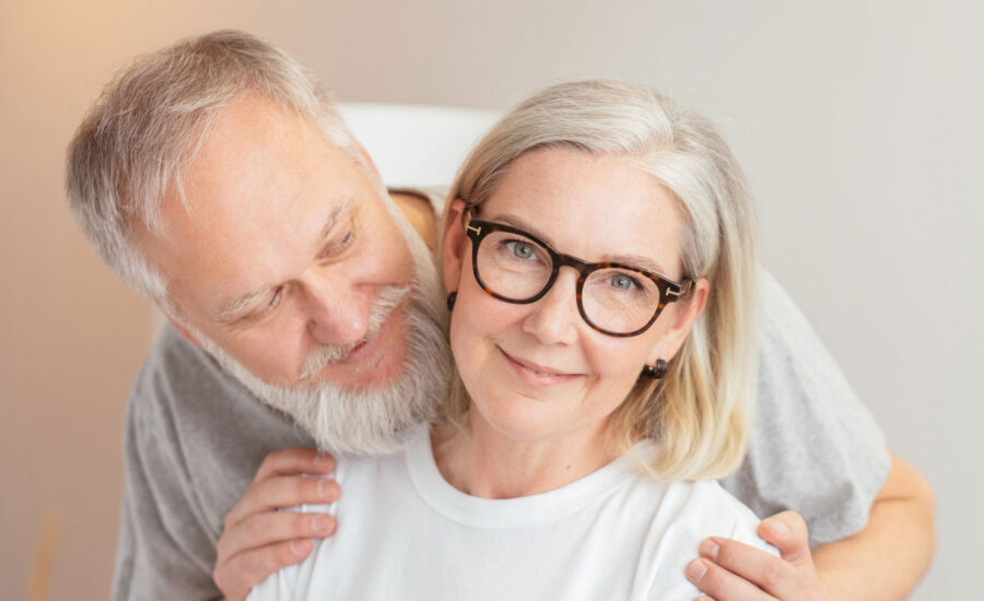 A woman in her late 50s/early 60s smiles, as her husband is behind her about to give her a hug. They're wondering about GICs for his retirement savigs